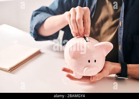 Primo piano di un uomo irriconoscibile seduto al tavolo e mettendo monete in piggybank, risparmiando denaro in crisi Foto Stock
