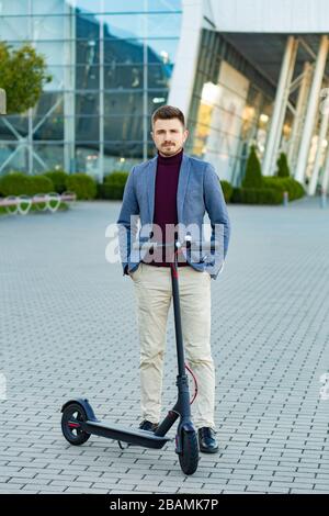 Giovane e bello uomo elegante con e-scooter in piedi sul marciapiede vicino al centro affari al tramonto. Trasporto urbano alla moda su moderno impianto elettrico Foto Stock