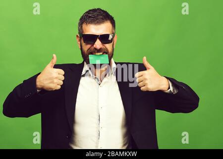 Ragazzo con felice viso e occhiali da sole isolato su sfondo verde. Concetto di business e successo. Uomo d'affari con carta vuota mostra pollici in su, copia spazio. L'uomo con la barba tiene il biglietto da visita nei denti Foto Stock