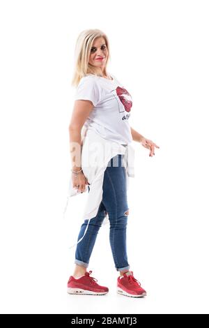 Elegante cool mezza età bionda capelli donna camminare e sorridere alla macchina fotografica indossando jeans. Corpo intero isolato su sfondo bianco. Foto Stock