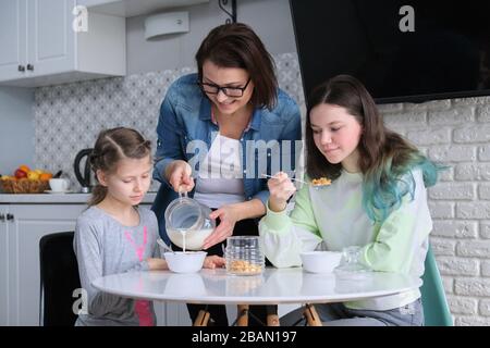 Madre felice che si prende cura di figlie in cucina. Mangiando le ragazze seduti al tavolo, colazione mattutina, latte in caraffa e fiocchi di mais dolci. Famiglia, mangiare, com Foto Stock