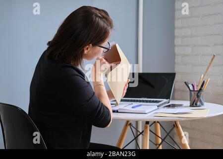 Donna matura seduto a casa alla scrivania con computer portatile, donna che guarda in una busta con documenti cartacei Foto Stock