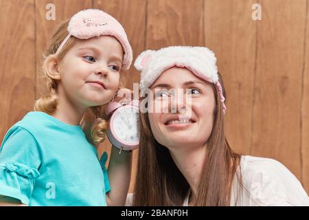 Mamma con una piccola figlia nel sonno maschera sbadiglio e abbracciarsi. Tenere un allarme vicino alla testa. È ora di dormire. Buona notte e buona mattina. Atto mattutino Foto Stock