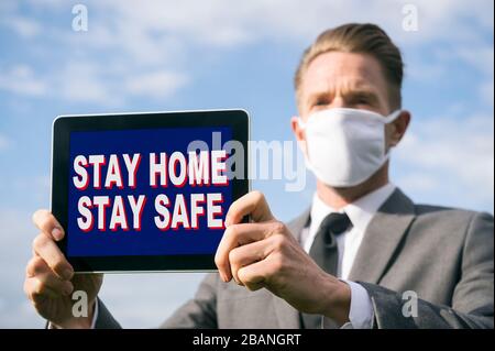 Uomo d'affari che indossa la maschera facciale protettiva COVID-19 tenendo un tablet con un Stay Home, Stay Safe message Foto Stock