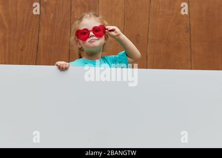 Piccoli occhiali a forma di cuore sorridenti ragazza bambino stando dietro un pannello bianco vuoto su sfondo di legno. Faccia divertente. Peeking fuori da dietro una ba Foto Stock