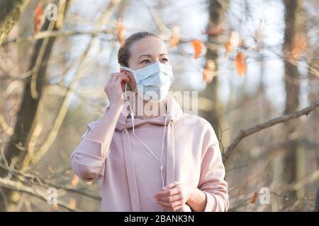 Ritratto della donna sportiva caucasica che indossa la maschera facciale di protezione medica mentre cammina nel parco, rilassandosi e ascoltando la musica. Corona virus, o Covid Foto Stock