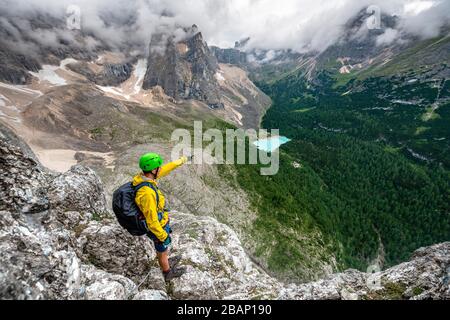 Giovane uomo punta in lontananza, alpinista su via ferrata Vanelli, vista sul Lago di Sorapis, circuito Sorapiss, montagne con nuvole basse Foto Stock