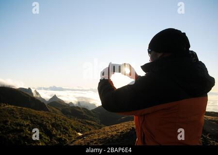 Alpinista scattando una foto al cellulare dell'alba a Pedra do Açú (2.245 m) nel Parco Nazionale Serra dos Órgãos, al punto più alto della città di Foto Stock