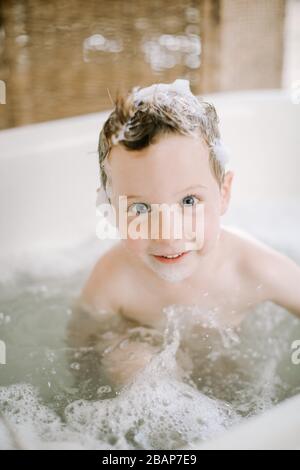 Ragazzo prescolare in vasca con bolle sul viso e capelli che guardano la macchina fotografica e sorridono Foto Stock