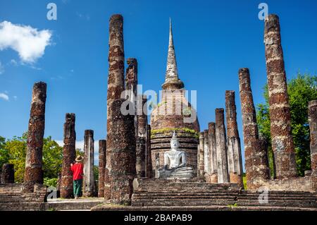 Tourist donna in costume rosso con fotocamera guardando antichi Buddha surround rovinato dalla colonna in Wat Sa Si di Sukhothai Historical Park, Thailandia Foto Stock