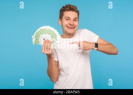 L'uomo eccitato overjoyed nella t-shirt bianca felice di tenere i soldi grandi, indicando le fatture dell'euro in mano e sorridente eccitatamente, godendo la vittoria inaspettata della lotteria. Foto Stock