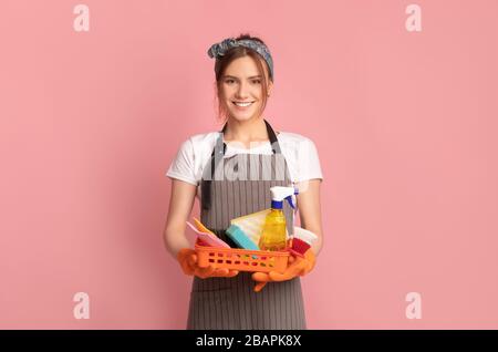 Donna sorridente in cesto Apron Holding con forniture per la pulizia, sfondo rosa Foto Stock