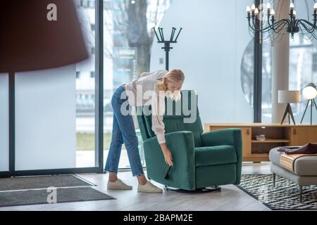 Poltrona grande e facile. Donna bella in jeans in piedi piegato da una morbida poltrona verde, testandolo, in un buon umore. Foto Stock