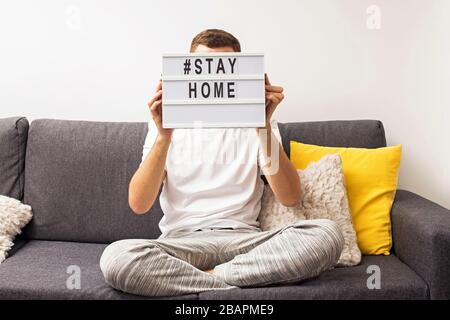 Uomo seduto sul divano nel suo soggiorno tenendo lightbox con messaggio Stay Home. Foto Stock