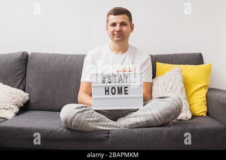 Uomo seduto sul divano nel suo soggiorno tenendo lightbox con messaggio Stay Home. Foto Stock