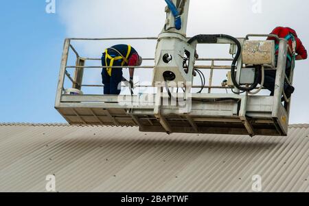 I lavoratori nella rampa di sollevamento idraulica riparano il gocciolatoio del tetto. Costruttori su piattaforma di sollevamento del braccio in piedi vicino alla parete dell'edificio, costruzione e riparazione w Foto Stock