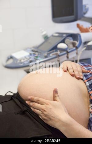 Pancia di donna incinta con apparecchio ultrasonografico sullo sfondo, femmina che tiene la pancia con le mani su di esso Foto Stock