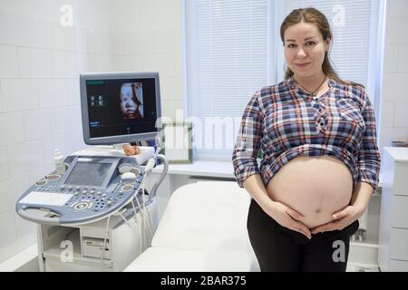 Felice incinta caucasica donna in piedi nella sala di consulenza dopo ultrasonografia vicino ecografia scansione con feto 3d immagine sullo schermo del monitor Foto Stock