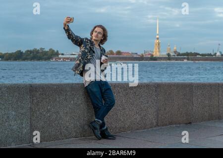 I bei uomini aspettano qualcuno, beve caffè e guarda al telefono, è vestito con una giacca militare e jeans, Pietro e Paolo fortezza è acceso Foto Stock