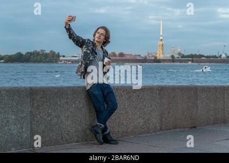 I bei uomini aspettano qualcuno, beve caffè e guarda al telefono, è vestito con una giacca militare e jeans, Pietro e Paolo fortezza è acceso Foto Stock