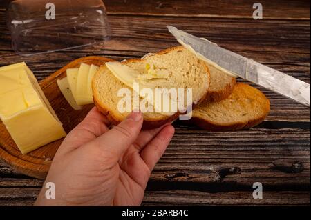 qualcuno spalma il burro su pane tostato di grano fresco con un coltello, un piatto di burro di legno con burro e fette di formaggio su uno sfondo di legno. Primo piano Foto Stock