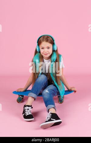 Bella bambina bionda piccola 7-10 anni in giacca in posa isolata su sfondo rosa pastello bambini ritratto. Concetto di stile di vita infantile. Simulazione dello spazio di copia. Tenere lo skateboard blu Foto Stock