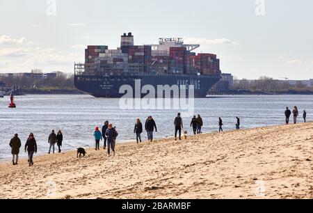 Amburgo, Germania. 29 marzo 2020. Gli escursionisti camminano lungo la spiaggia dell'Elba Övelgönne, mentre sullo sfondo la nave container YM wellhead sta navigando a valle. Credit: Georg Wendt/dpa/Alamy Live News Foto Stock