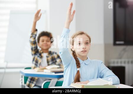 Ritratto di ragazza bionda carino che alza la mano per rispondere mentre si siede alla scrivania in classe scuola, copia spazio Foto Stock