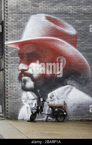 Opere d'arte su larga scala su una parete di un edificio raffigurante un uomo che indossa un cappello da cowboy rosa. Accanto a questo è parcheggiato uno scooter Foto Stock