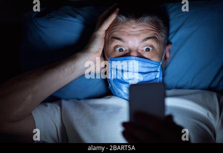 Uomo spaventato nella maschera usando lo smartphone che giace nel letto all'interno Foto Stock