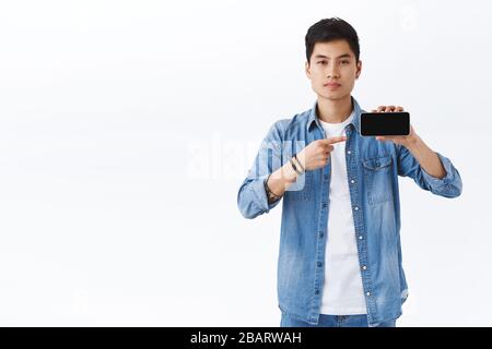 Tecnologia, concetto di stile di vita online. Ritratto di uomo asiatico sicuro e di buon aspetto che mostra app o promo dell'azienda sullo schermo dello smartphone, indicando Foto Stock