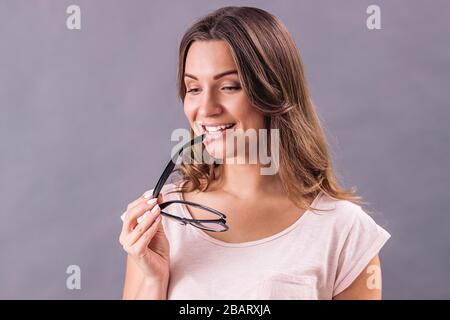 Ritratto di giovane bella donna che tiene occhiali in mano e sorridente mentre si sta isolati su sfondo grigio. Felice ragazza attraente in T-s rosa Foto Stock