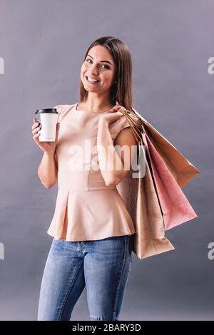 Felice bella donna che tiene una tazza di caffè in una mano e borse per lo shopping in un altro mentre isolato su sfondo grigio. Ragazza attraente bere cof Foto Stock