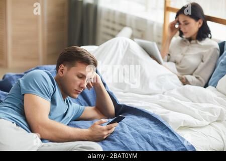 Ritratto di giovane coppia moderna utilizzando dispositivi mobili mentre si siede sul letto, comunicazione problema concetto, copia spazio Foto Stock