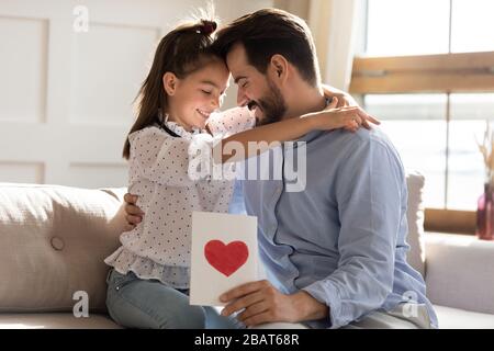 Un giovane padre grato che tiene un biglietto d'auguri fatto a mano, una figlia coccolante. Foto Stock