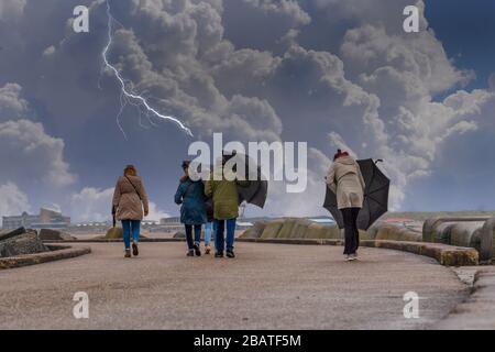 Un gruppo di persone che camminano e cercando di mantenere gli ombrelli in una giornata di vento e fulmini in background Foto Stock