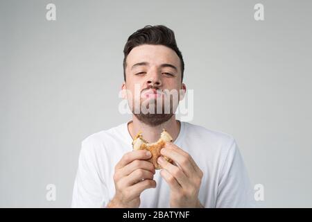 Giovane uomo isolato sullo sfondo. Buon ragazzo soddisfatto guardare su macchina fotografica e hamburger da masticare. Mangiare cibo cattivo fatning con piacere. Digiuno nelle mani. Foto Stock