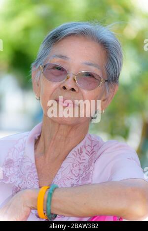 Testa colpo di primo piano faccia di una bella donna sana anziani asiatici vestito in stile tailandese abiti, ritratto vecchia signora rilassante all'aperto nel Foto Stock