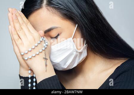 Una donna che indossa una maschera medica che prega sul coronavirus pandemia globale per ridurre l'ansia e la depressione. Donna che tiene il rosario. Foto Stock