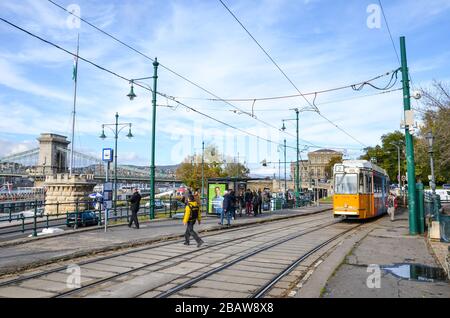 Budapest, Ungheria - 6 novembre 2019: Tram giallo alla stazione pubblica. Persone che camminano intorno. Il ponte della catena Szechenyi, l'edificio MTA e il centro storico sullo sfondo. Persone sulla strada. Giornata di sole. Foto Stock