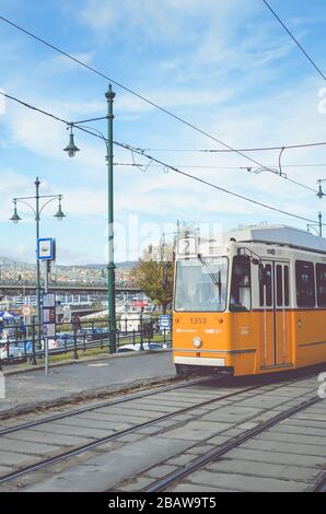 Budapest, Ungheria - 6 novembre 2019: Tram giallo in una stazione del tram. Il ponte della catena Szechenyi e il centro storico sullo sfondo. Trasporto pubblico ungherese. Foto verticale con filtro. Foto Stock