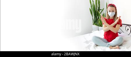 Una giovane donna bionda è seduta su un letto bianco durante il periodo di quarantena a causa del virus COVID-19, indossa una maglietta rossa, confortevole h. Foto Stock
