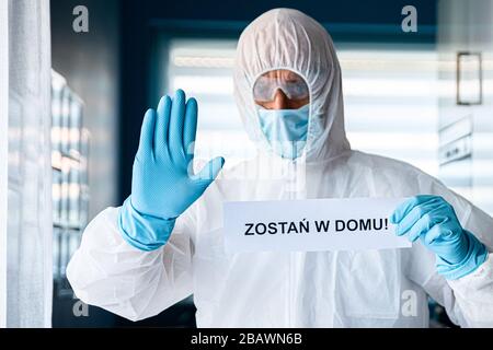 Uomo in tuta anti virus protettiva e maschera con scheda di tenuta con messaggio rimanere a casa in lingua polacca Foto Stock