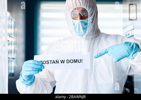 Uomo in tuta anti virus protettiva e maschera con scheda di tenuta con messaggio rimanere a casa in lingua polacca Foto Stock