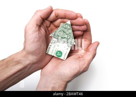 La fattura americana del dollaro piegata nella forma di una casa di origami seduta cullata in un paio di mani contro uno sfondo bianco Foto Stock