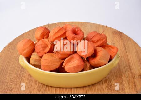 Physalis rosso commestibile in recipiente in ceramica gialla su tagliere in legno su sfondo bianco Foto Stock