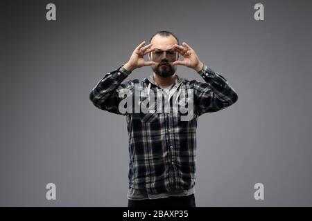 Uomo con bearded in camicia in piedi e tenendo un pezzo di vetro colorato trasparente davanti ai suoi occhi in entrambe le mani, guardandolo attraverso la fotocamera. Foto Stock