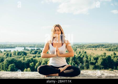 Giovane donna pratica yoga fuori. Ragazza sorridente calma seduta sul parapetto in posizione lotus . Le sue mani a livello del torace si toccano. Alberi fiume Foto Stock