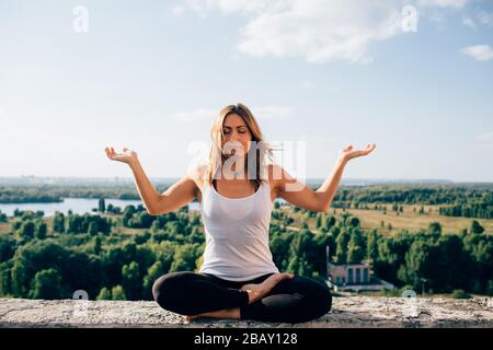 Giovane donna pratica yoga fuori. Ragazza bionda calma seduta sul parapetto in posizione lotus con occhi chiusi. Le sue palme si voltarono verso il cielo. Alberi fiume e. Foto Stock