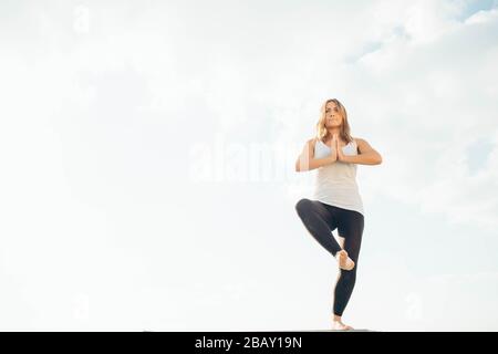 Giovane donna pratica yoga fuori. Bionda ragazza in piedi su una gamba, un'altra sollevata piegata al ginocchio. Mani a livello del torace, palmi che toccano. Cielo e nuvole Foto Stock
